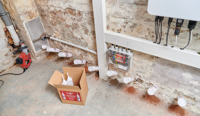 Isofin®-Flaschen stecken in einer Bohrlochreihe am Sockel einer Kellerwand, daneben Karton und Bohrhammer